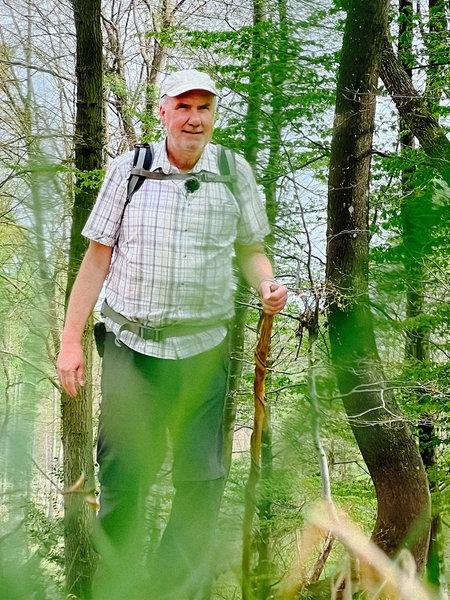 Ralf Spilles, ein Mann mit Kappe und leichtem weißem Bart und Gehstock in der Hand. Im Hintergrund ein dicht bewachsenener Wald.