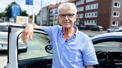 Uwe Blaschke steht vor einem Auto. Er trägt ein blaues Hemd und eine Brille.