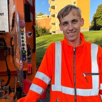 Laurin Oppermann steht im orangenen Overall vor einem Müllfahrzeug.
