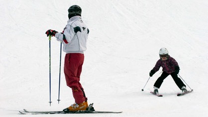 Auf der Skipiste vom Hesselbacher Gletscher steht eine erwachsene Person mit Ski und Ski-Klamotten und blickt nach hinten zu einem kleinen Mädchen, das ebenfalls Ski trägt und gerade bremst.