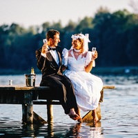 Ein Brautpaar sitzt auf einem Steg am Wasser und hält ein Sektglas in der Hand.