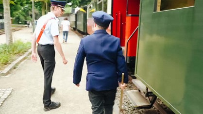 Leo läuft, begleitet von einem Schaffner, mit einem Hammer in der Hand und in Uniform an der roten Selfkantbahn entlang.