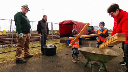 Drei Erwachsene und zwei Kinder stehen vor einer Halle, im Hintergrund sind Bahngleise zu sehen