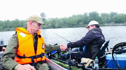 Johannes Pantel mit beiger Cap und orangener Weste reicht einem Mann im Nachbarboot die Hand und zieht ihn ans Boot heran.