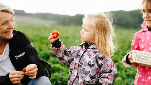 Ein kleines Mädchen hält eine rote Erdbeere in der Hand. Eine Frau lächelt das Mädchen an.