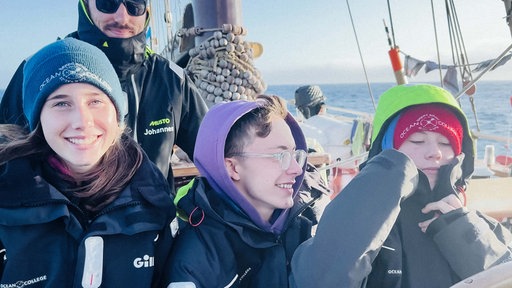 Mehrere Personen befinden sich auf einem Schiff. Im Vodergrund stehen drei Jugendliche. Sie lächeln. Hinter ihnen steht ein Mann. Er lächelt ebenfalls.