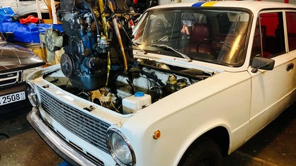 Ein weißer Lada steht in einer Werkstatt. Die Motorhaube ist geöffnet. Der alte Verbrennungsmotor wurde gegen einen Elektromotor ausgetauscht und die neue Elektrik wird verkabelt.