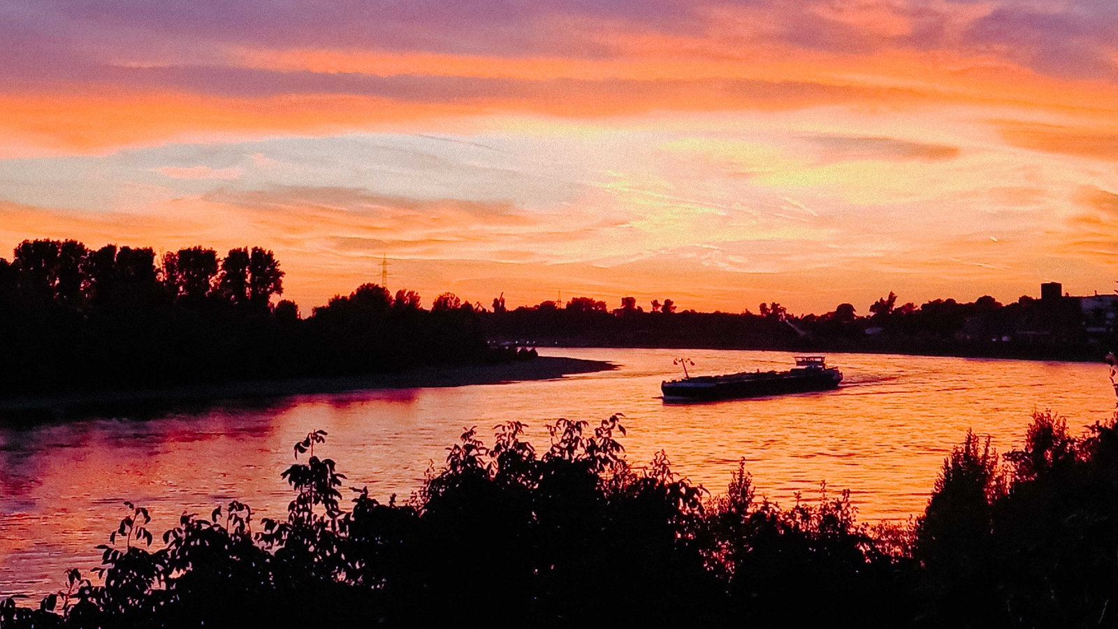 Sonnenuntergang über einem großen Fluss, auf dem eine kleine Fähre fährt. 