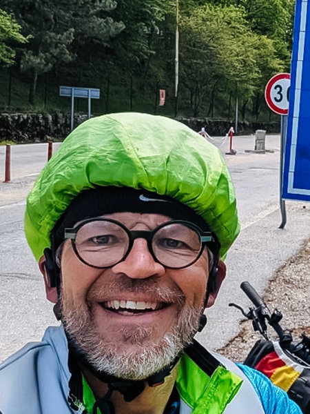 Rainer Stephan, Brille, grauer Bart und grüner Regenschutz über dem Helm, steht vor einem Landesschild der Türkei und zeigt lächelnd einen Daumen hoch in die Kamera