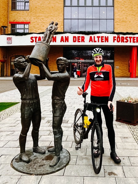 Frank Müller steht mit seinem Rad im Radtrikot vor dem Fußballstadion "Alte Försterei" von Union Berlin neben einer Statue von zwei Spielern, die einen Pokal hochhalten