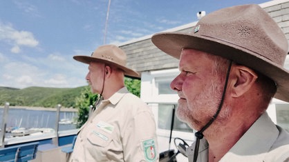 Zwei Männer schauen nach links auf einen See. Sie tragen beige Kleidung und Ranger-Hüte.