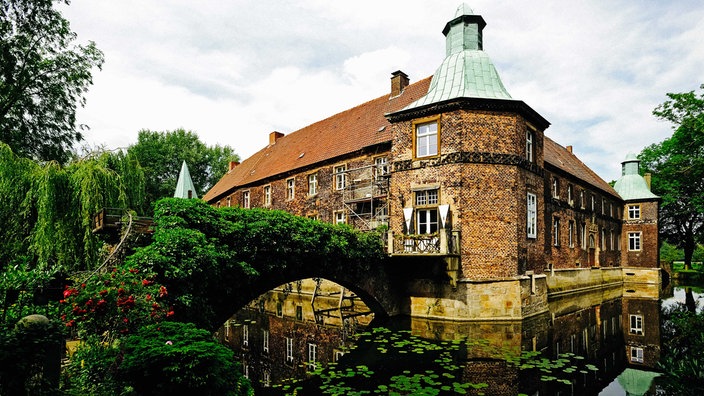 Renaissance-Schloss aus hellbraunem Backstein und grün-weißen Türmchen, umgeben von einem Wassergraben und einer grün-überwucherten Steinbrücke