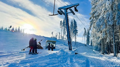 Eine verschneite Winterlandschaft mit einem Skilift, mehreren Menschen auf Skiern, hell leuchtender Sonne und schneebedeckten Bäumen am Waldrand.