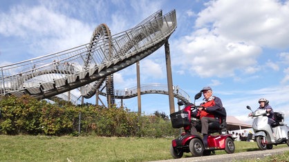 Zwei ältere Menschen mit Elektromobilen fahren vor der Skulptur "Tiger and Turtle" in Duisburg.