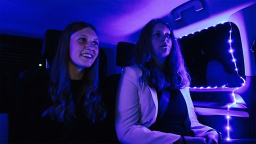 Zwei junge Mädchen sitzen im Party-Shuttle. Es ist dunkel, blaue LED-Lichter leuchten.