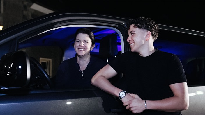 Kerstin Schulz-Höfel sitzt im Party-Shuttle und schaut aus dem Fenster. Neben dem Fenster steht ihr Sohn Sam. Beide lächeln.
