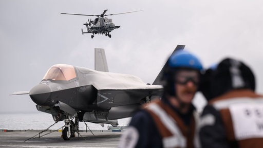 Personen stehen vor Kampfjet, ein Hubschauber fliegt über ihnen 