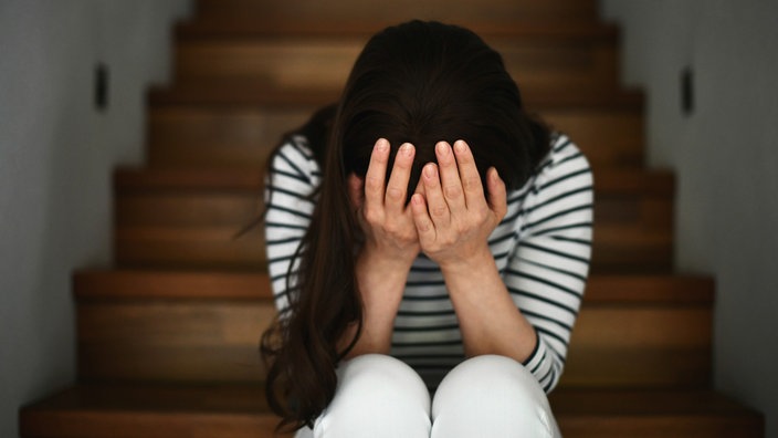 Das Beitragsbild des Dok5 "Erschütterungen – über eine Mutter, die ihr Kind tötet" zeigt eine verzweifelte Frau auf der Treppe sitzend den Kopf in die Hände gelegt. 