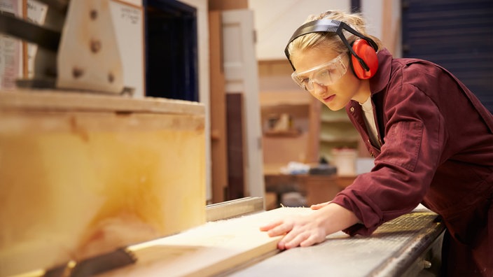 Das Beitragsbild des Dok5 "Lost in Transition" zeigt eine Auszubildende in Schutzkleidung und Schutzbrille an einer Holzsäge.