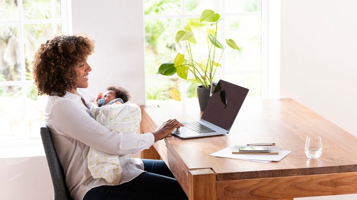 Das Beitragsbild des Dok 5 "Mütter im Job - Vereinbarkeit von Familie und Beruf" zeigt eine Mutter, die mit ihrem Säugling im Arm am Esstisch sitzt und am Laptop arbeitet. 