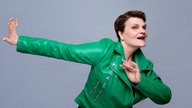 Gayle Tufts am Tanzen: Ein Arm ist zur Seite gestreckt, der andere vor ihrem Oberkörper gehalten. Sie trägt eine lechtende, grüne Lederjacke und roten Lippenstift.