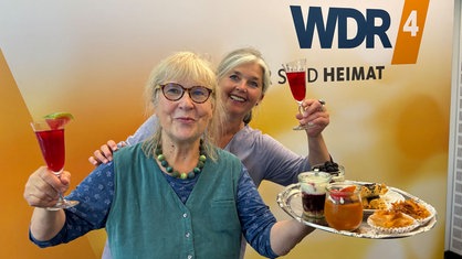 Ulla Scholz und Heike Knispel im WDR4-Studio. Beide lächeln einladend in die Kamera und halten Sektgläser hoch. Ulla hält außerdem ein Tablett mit verschiedenen Speisen. 