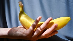 Eine Frauenhand hält eine Banane, im Hintergrund hängt ein blauer Vorhang