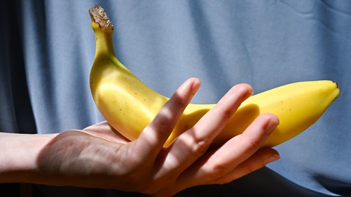 Eine Frauenhand hält eine Banane, im Hintergrund hängt ein blauer Vorhang