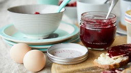 Ein Frühstückstisch mit Eiern, einem Marmeladenbrot und dem Marmeladenglas sowie einer Schüssel Müsli im Hintergrund. 
