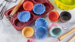 Von oben fotografiert: verschiedene Backutensilien verstreut auf einer Arbeitsfläche, darunter ein Muffinblech und viele bunte Muffinformen aus Silikon.
