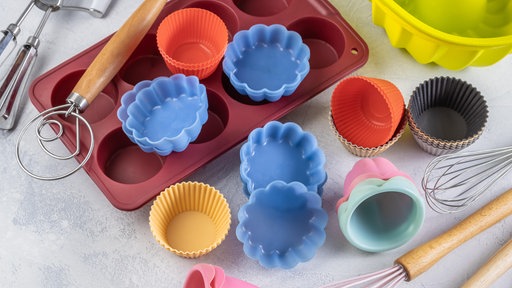 Von oben fotografiert: verschiedene Backutensilien verstreut auf einer Arbeitsfläche, darunter ein Muffinblech und viele bunte Muffinformen aus Silikon.
