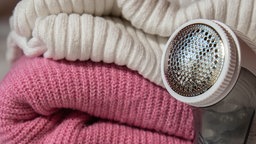 Zwei Pullover in weiß und rosa liegen gefalten aufeinander, neben ihnen ein Fussel-Rasierer