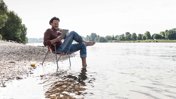 Symbolbild: Ein Mensch sitzt lächelnd mit einem Tablet alleine auf einem Campingstuhl am Wasser.