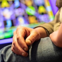 Hände eines Mannes liegen auf seinem Schoß. Im Hintergrund ist ein Spielautomat zu sehen.