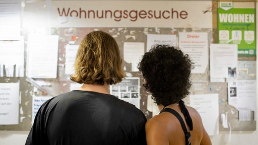 Ein Paar steht vor einer Anzeigentafel mit der Aufschrift "Wohnungsgesuche" an der Uni in Bonn.