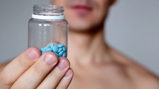 Ein junger Mann hält eine Plastikdose mit blauen Viagra-Tabletten in die Kamera