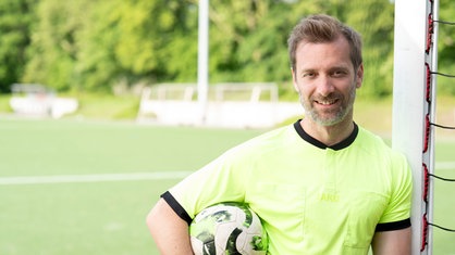 Daniel Aßmann ist an einem Tor angelehnt und hat einen Fußball unter dem Arm. 
