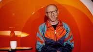 Ein Mann mit Brille und Glatze in bunter Trainingsjacke vor rotem Hintergrund