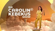 Carolin Kebekus grinst und hebt den rechte Zeigefinger gestreckt nach oben. Sie ragt aus bunten neon Leuchtringen heraus. 
