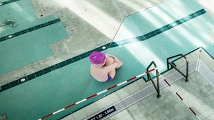 Ein Mädchen in Schwimmbekleidung sitz kauernd am Beckenrand, den Kopf auf die Knie gesenkt, und umschlingt die Beine mit den Armen.