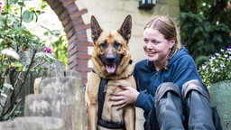 Das Bild zeigt einen Schäferhund (links) und eine junge Frau (rechts), die zusammen auf einer Treppe sitzen.