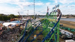 Menschen sitzen in einem Wagen auf einer Achterbahn bei der Rheinkirmes Düsseldorf. Im Hintergrund ist der Rhein zu sehen.