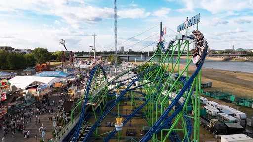 Menschen sitzen in einem Wagen auf einer Achterbahn bei der Rheinkirmes Düsseldorf. Im Hintergrund ist der Rhein zu sehen.