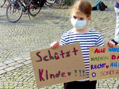 Ein Kind mit Mundschutz bei einer Kundgebung: Das Mädchen hält zwei Plakate hoch mit den Aufschriften "Schützt Kinder" und "Alle Kinder haben das Recht auf Schutz"
