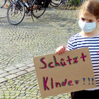 Ein Kind mit Mundschutz bei einer Kundgebung: Das Mädchen hält zwei Plakate hoch mit den Aufschriften "Schützt Kinder" und "Alle Kinder haben das Recht auf Schutz"