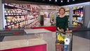 Anna Planken mit einem vollen Einkaufswagen in einem Studio. Im Hintergrund ist ein Bild von gefüllten Regalen in einem Supermarkt.