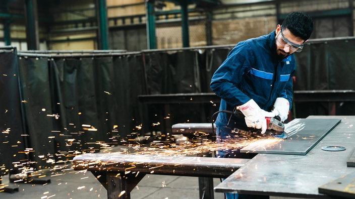 Ein Azubi mit Schutzbrille schleift eine Metallplatte, dabei fliegen viele Funken