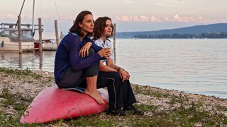 Zwei Frauen sitzen auf einem umgedrehten Surfboard am Strand und schauen auf den See.