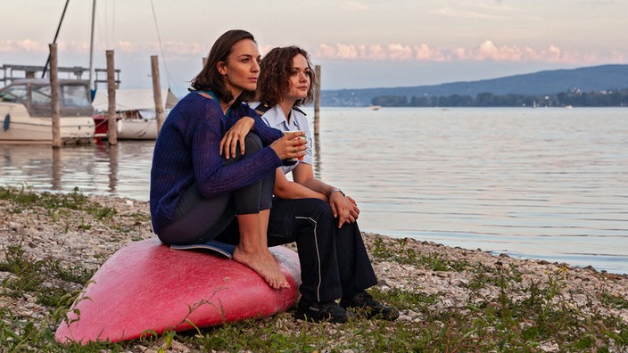 Zwei Frauen sitzen auf einem umgedrehten Surfboard am Strand und schauen auf den See.