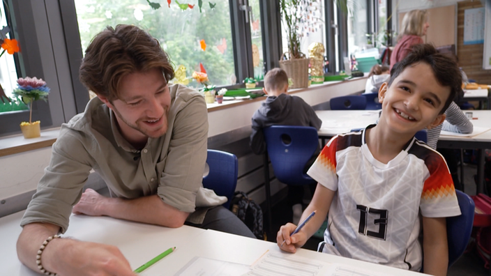 Ein junger Schüler sitzt mit einem Reporter an einem Tisch in der Schulklasse, sie halten je einen Stift in der Hand und bearbeiten gemeinsam Aufgaben